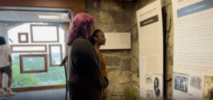 Two women stand viewing an exhibit, a window viewing the mountains is in the background