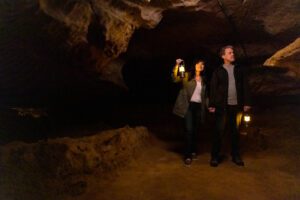 A male and female couple walk in a dimly-lit cavern holding lanterns.