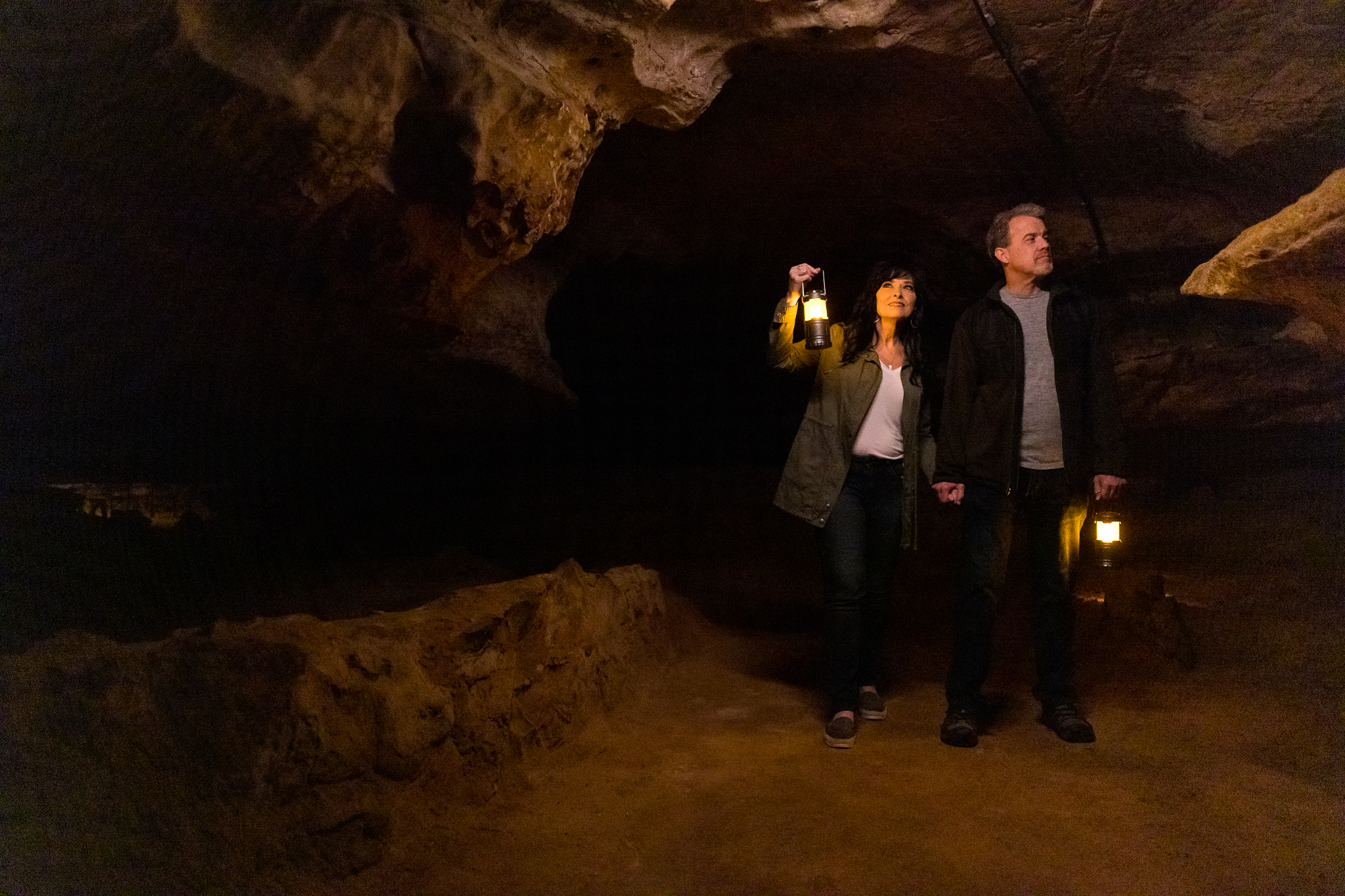 A male and female couple walk in a dimly-lit cavern holding lanterns.