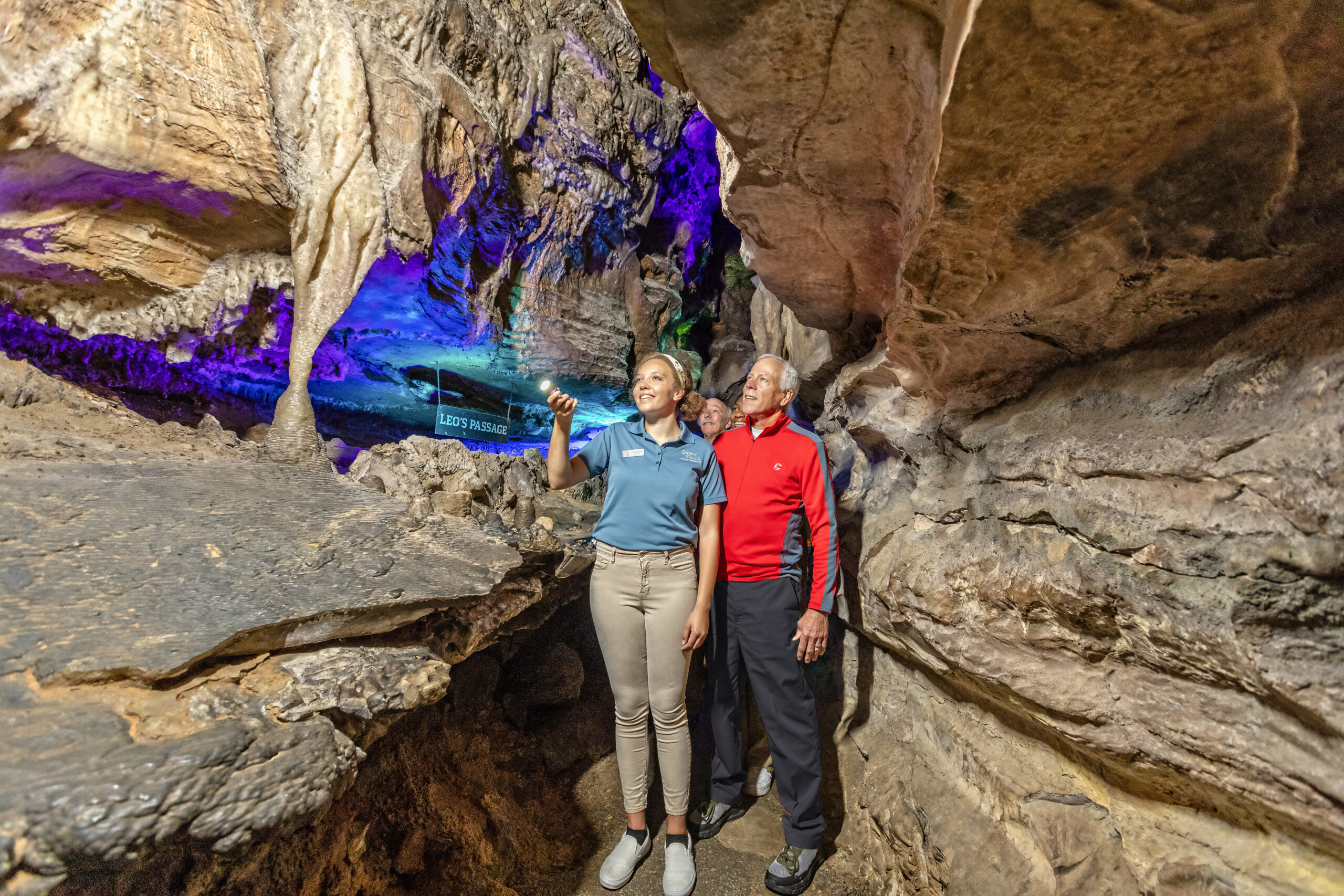 A tour guide in a blue shirt shines a light into the caves for a group of visitors behind her. A man in a red shirt stands next to her, looking at the caves