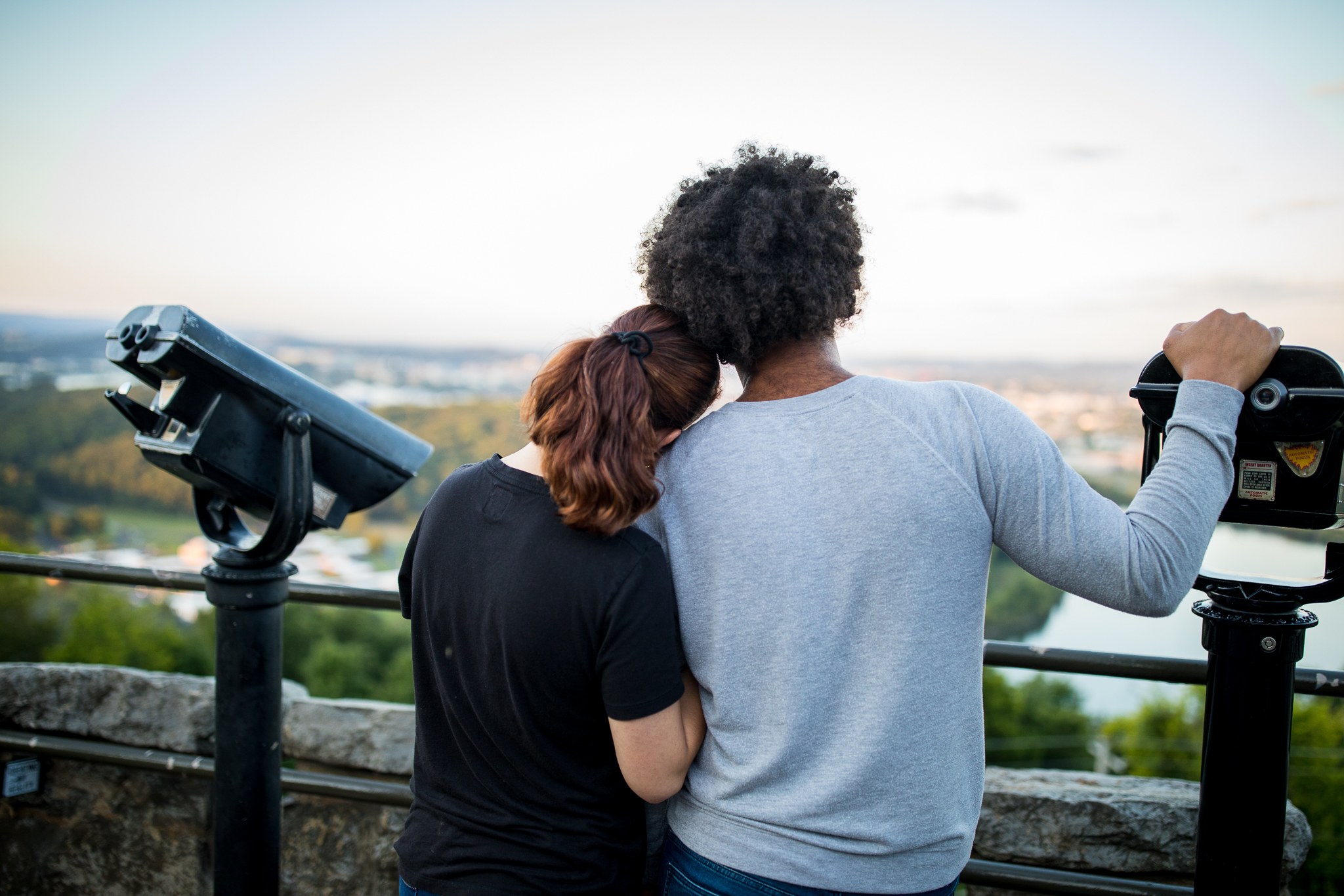 A male and female multi-racial couple stand on the top of the tower lookout with their bakcs to the camera, enjoying the view. The woman rests her head on the man's shoulder.