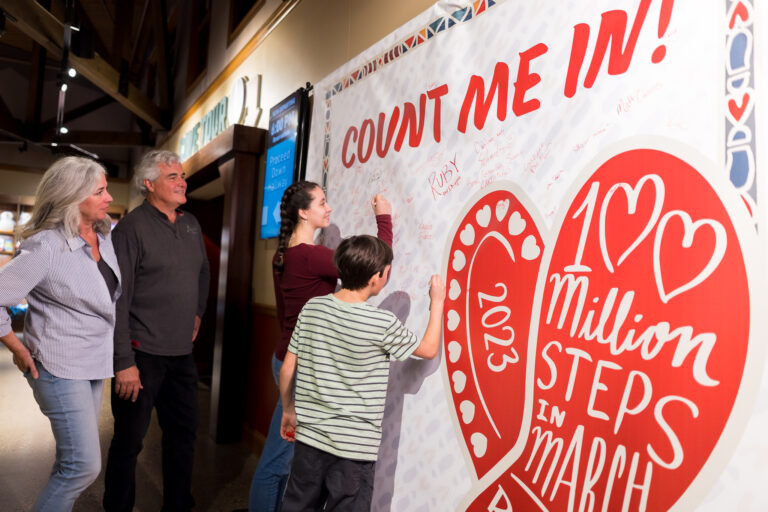 Man and woman, stand indoors in front of large banner while teenage girl and tween boy sign red and white banner showing their participation in 100 Million Steps in March.