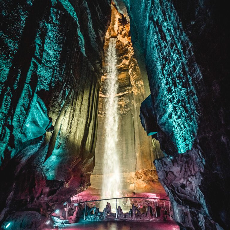 Photo of the Ruby Falls waterfall inside the cave. The cave walls are lit with teal, gold, and magenta colored LED lighting