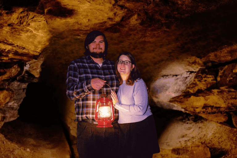 A tall man stands next to a woman inside a limestone cave with one arm around the woman while the other hand holds a lit red lantern in front of his body.