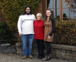 Two Steiner Scholarship winners are pictured in front of a low stone wall with Motiva Steiner in the middle wearing a red sweater and black pants. Man on left is wearing a white sweater with a black stripe and jeans. On the left, a woman wears a brown dress.