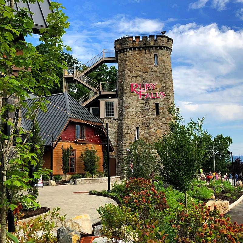 Summertime view of the Village Plaza and historic Lookout Mountain Tower at Ruby Falls. Landscaping surrounds the plaza and a group of visitors are walking down the ramp near the tower.