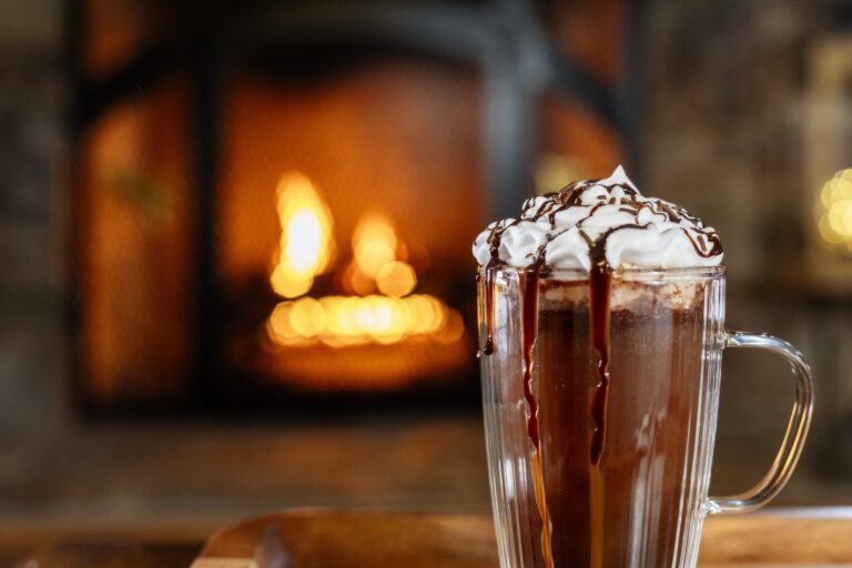 Hot chocolate in a glass mug with whipped cream on the top drizzled with chocolate sauce is placed in front of a fire in a fireplace.