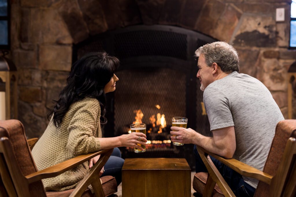 Middle age man and woman sitting in front of the stone fireplace drinking craft beer in pint glasses.