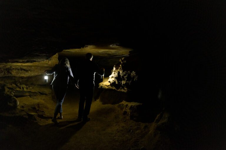 Man and woman with their backs to the camera walk on a cavern trail holding lanterns to light their way.