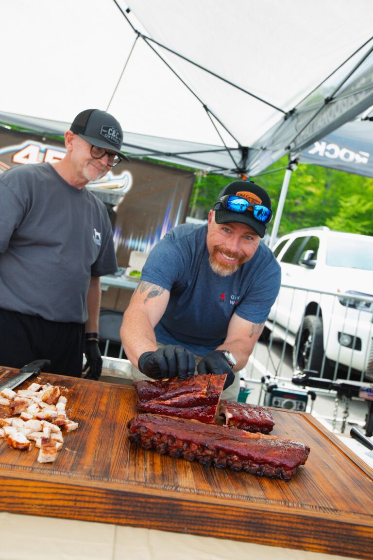 Man smiles as he crouches behind a wood table holding up a slab of ribs. A grill is behind the man and another man stands next to the smiling man.