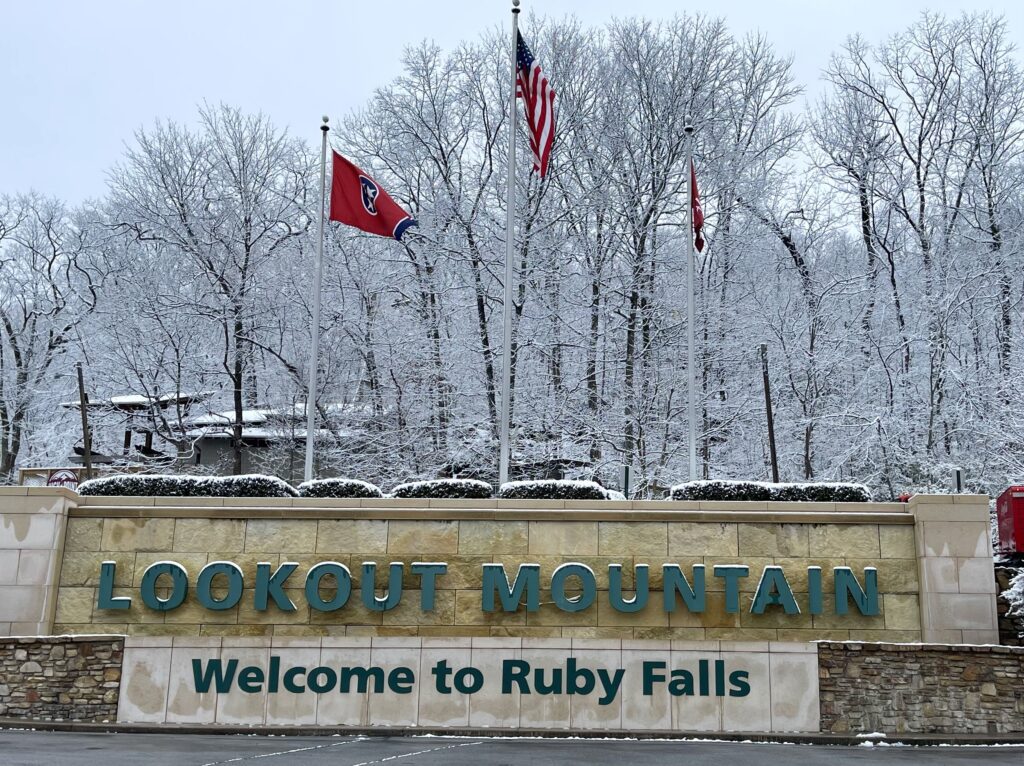 Trees are covered in snow behind a large stone entrance to Ruby Falls with 3 flags - flags for the United States, Tennessee and Ruby Falls