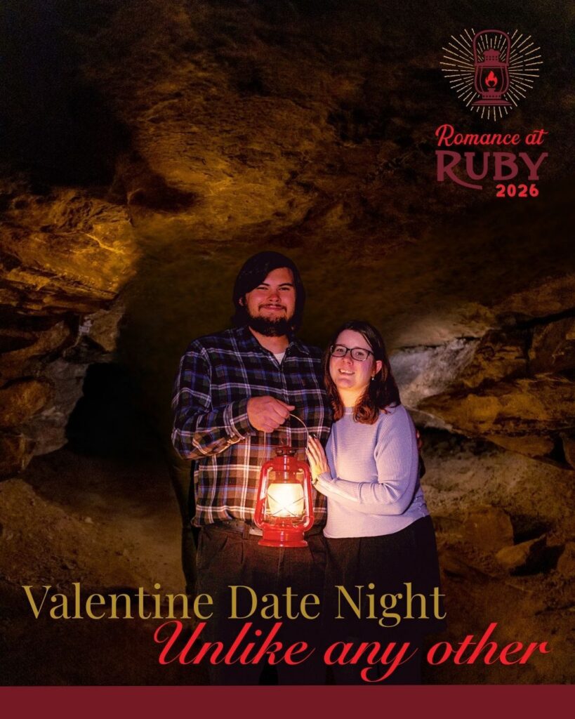 A man with dark hair and a bread stands in a cave with his arm around a women with dark hair and glasses while holding a red lantern.